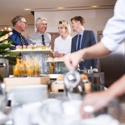 Distant four smiling business people chatting and standing at buffet table with food and blurred crockery and waiter in foreground
